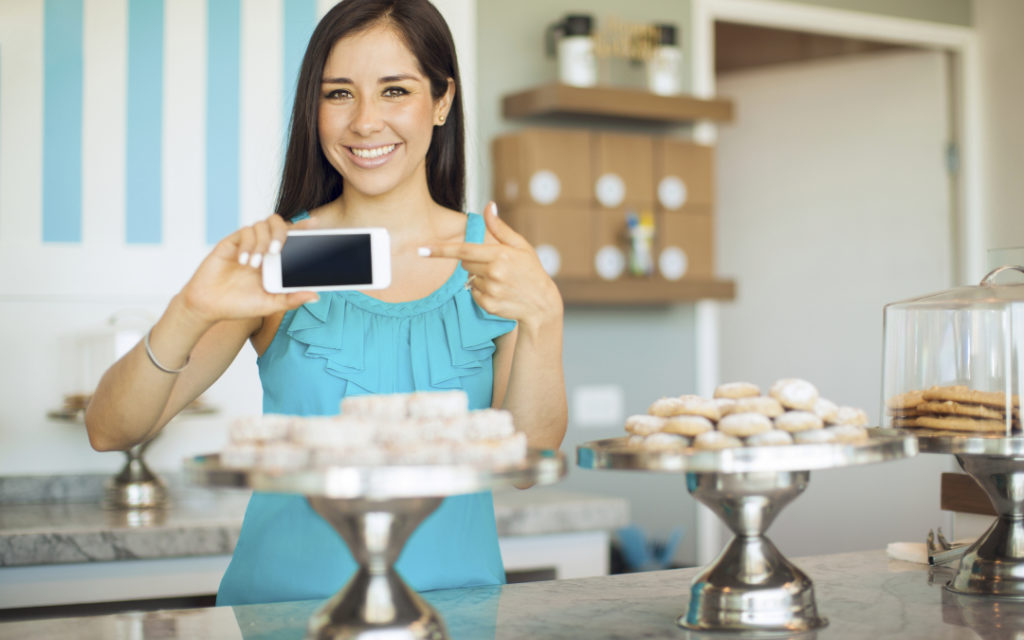 Pretty Hispanic young woman pointing at her smartphone screen and smiling in a bakery