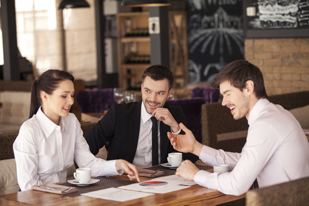 Three friends discuss graph lying on table at cafe. Business people having meeting at coffee table, smiling.