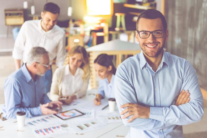 Handsome businessman in eyeglasses is looking at camera and smiling, in the background his colleagues are discussing work